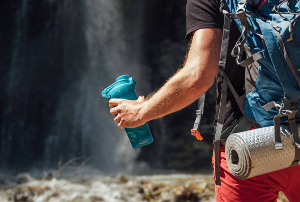 chiudi a mano con una bottiglia d'acqua potabile. uomo con zaino vestito con abiti da trekking attivi turistico stare vicino alla cascata del fiume di montagna e godersi la natura. immagine concettuale di viaggio e trekking - number of people human gender people waterfall foto e immagini stock