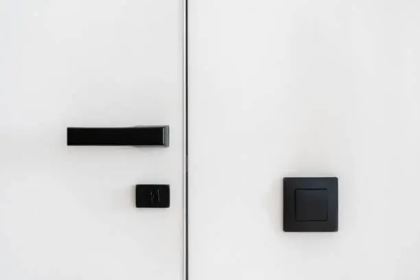 Modern choice for your home. Closeup of minimalistic black matt door handle with safety lock on closed white door and light switch on white wall in hotel room or contemporary apartment