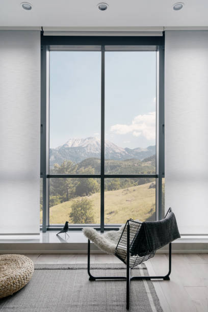 Vertical shot of modern metal chair on gray carpeted floor in front of large window with amazing mountain landscape in light coloured living room. Minimalist interior design in hotel room or house