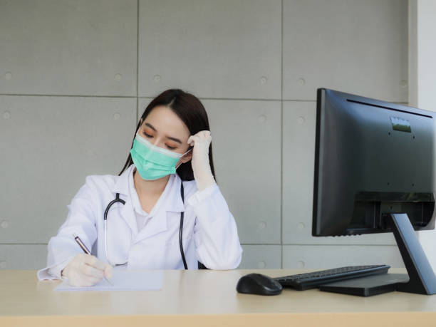 A female doctor or nurse wearing a surgical mask and medical gloves is sitting in a worrying mood, and her using a pen to writing information on the paper is on the desk. stock photo