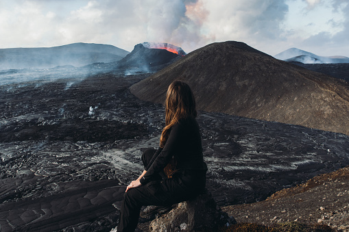 Scenic view of a woman explorer with long hair sitting at the edge of the mountain looking at the new volcano and the eruption site with lava in Iceland