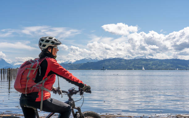 woman with electric mountain bike at Lake of Constance, Germany happy, active senior woman admiring a awesome view over Lake Constance with snow capped Swiss mountains in background bodensee stock pictures, royalty-free photos & images
