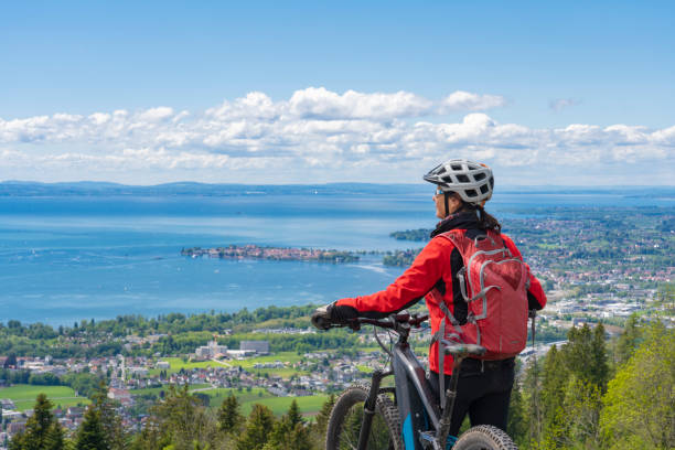 woman with electric mountain bike at Lake of Constance, Germany happy, active senior woman admiring a awesome view over Lake Constance with snow capped Swiss mountains in background bodensee stock pictures, royalty-free photos & images
