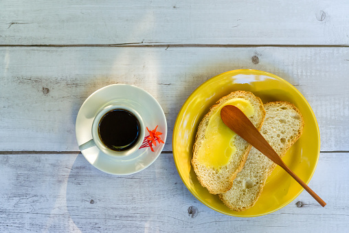 Ghee butter on sliced bread, with wooden spatula, in a yellow plate next to a cup of coffee, on a white wooden table.