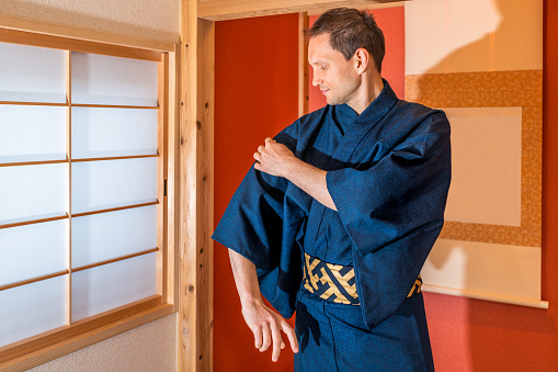 Traditional japanese machiya house room with window and red alcove with hanging scroll and man in blue kimono standing fixing clothes yukata at tea ceremony