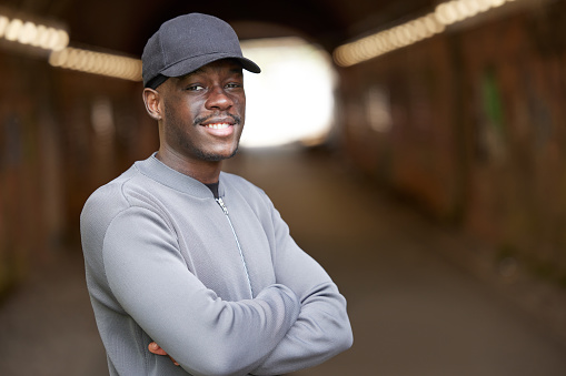 Portrait Of Smiling Young Man Wearing Baseball Cap Standing In Tunnel In In Urban Setting