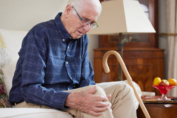 Senior Man Sitting On Sofa At Home Suffering With Knee Pain From Arthritis Senior Man Sitting On Sofa At Home Suffering With Knee Pain From Arthritis artificial knee photos stock pictures, royalty-free photos & images