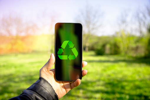 Mobile Phone with recycling symbol in the female hand against green nature. Recycling concept. Copy space