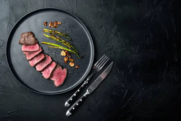 Juicy steak medium rare beef Filet Mignon or Eye Fillet set, with onion and asparagus, on plate, with meat knife and fork, on black stone background, top view flat lay, with copy space for text