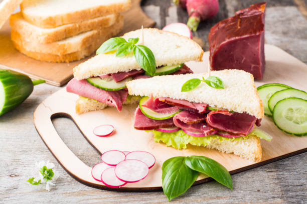 istock Fresh white bread sandwiches with pastrami, cucumber, radish and basil on a cutting board. American snack. Rustic style 1322562009