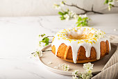 Fresh homemade lemon cake decorated with white glaze and zest on white marble background with branches of blossoming plum