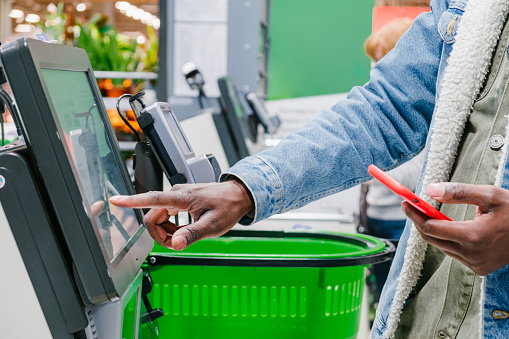 The finger of an African man in close-up at the supermarket checkout selects the desired product on the electronic screen of the cash register with a phone in his hands against the background of green shopping baskets, retail and self-service checkout in a hypermarket