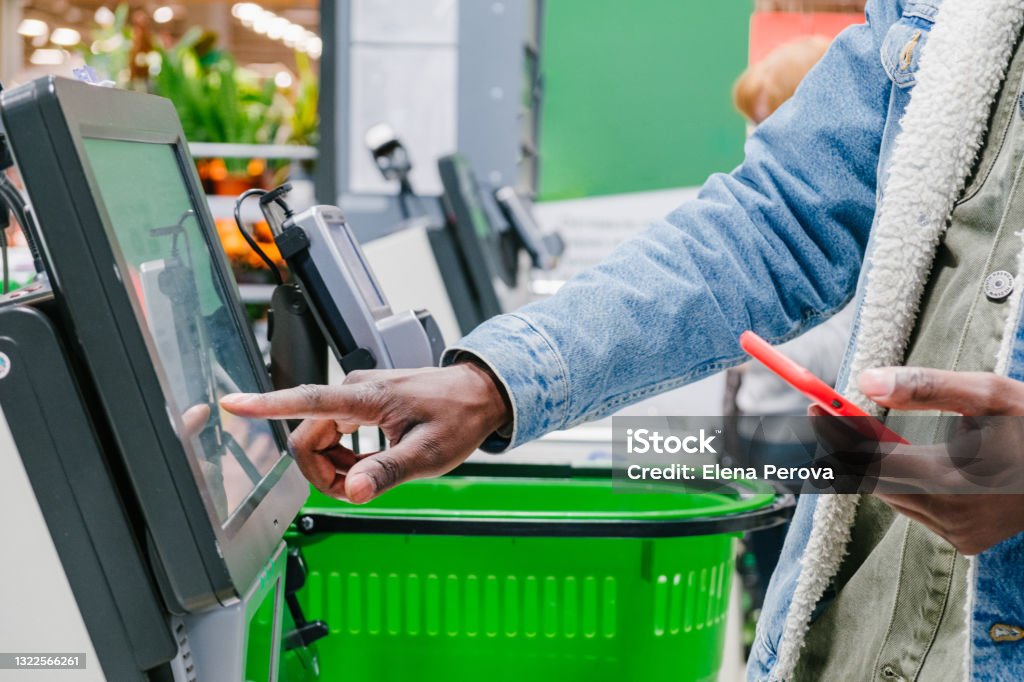 The finger of an African man in close-up at the supermarket checkout selects the desired product on the electronic screen of the cash register with a phone in his hands The finger of an African man in close-up at the supermarket checkout selects the desired product on the electronic screen of the cash register with a phone in his hands against the background of green shopping baskets, retail and self-service checkout in a hypermarket Supermarket Stock Photo