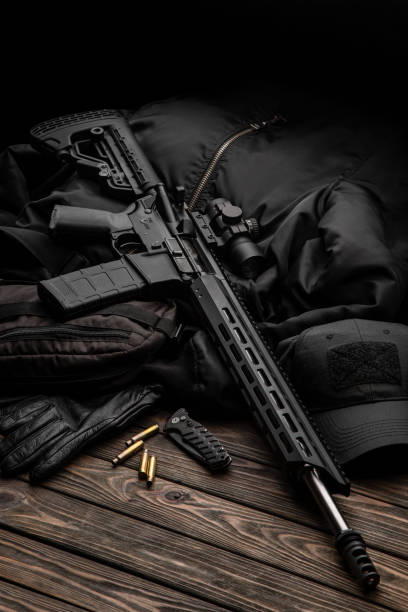 istock Modern automatic rifle with a telescopic sight on a dark background. Jacket, cap, gloves on a wooden table. The uniform of a guard or a mercenary. 1322568304