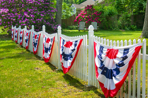 Two wooden Adirondack chairs rest under a canopy of colorful rhododendron bushes in a yard behind a white fence festooned with red, white and blue bunting  in anticipation of the upcoming Independence Day Holiday