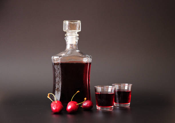 Cherry liqueur in a glass bottle and two glasses, next to ripe berries on a black background. Cherry liqueur in a glass bottle and two glasses, next to ripe berries on a black background. Close-up. cherry colored stock pictures, royalty-free photos & images