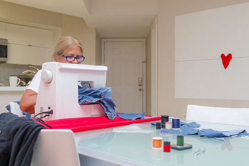 Portrait of blond mature latin female dressmaker working on a sewing machine in her design studio at home in United States of America USA, fixing clothes and jeans. She is wearing a white t-shirt.