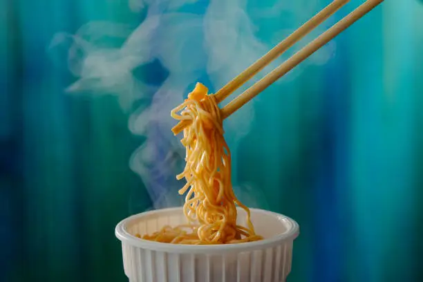 The most popular Asian fast food, available internationally, here a mouthful of freshly prepared with added boiling water instant cup noodles are on chopsticks, ready to be eaten. Scene set against a vibrant contrast turquoise-colored wood background. Shallow depth of field with the focus being on the noodles at the end of the bamboo disposable chopsticks.