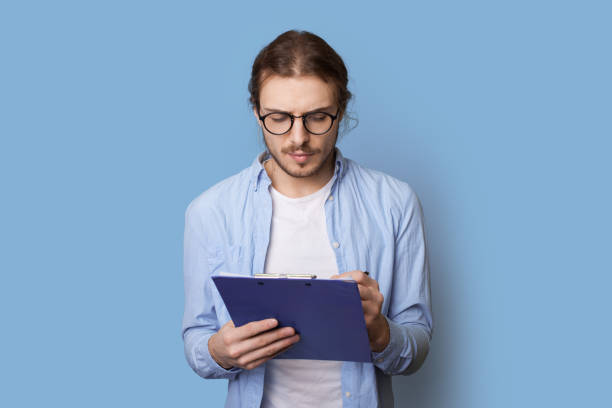 istock Concentrated man with long hair and beard is writing something in a folder on a blue studio wall 1322623782