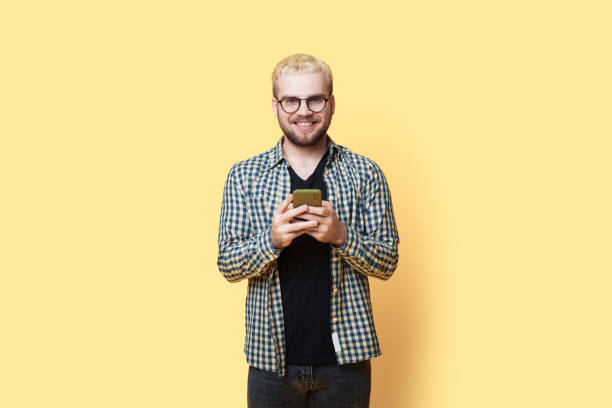 istock Blonde bearded man with glasses is smiling at camera while chatting on a mobile posing on a yellow studio wall 1322623808
