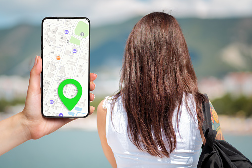 A females hand holds a smartphone with an online map app and a marked destination. In the background, a woman looks at the sea, a view from the back. Concept of online navigation and modern technologies.
