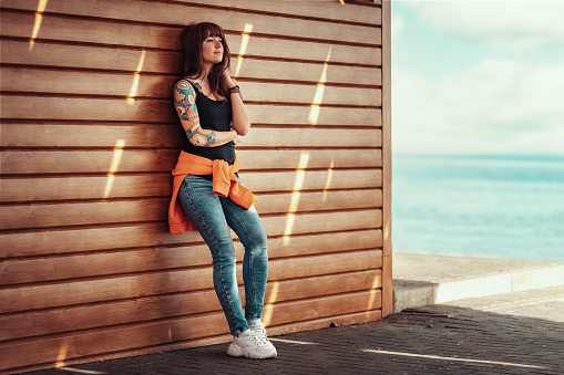 A young beautiful woman with tattoos on her arm, posing near a wooden wall. Sea and beach on the background.