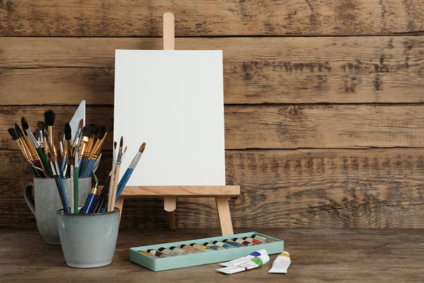 istock Easel with blank canvas, paints and brushes on wooden table. Space for text 1322640387