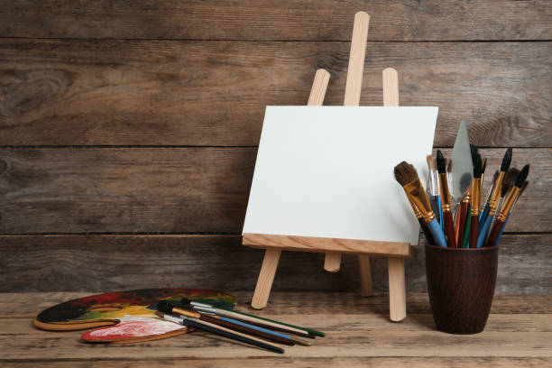 istock Easel with blank canvas, palette and brushes on wooden table 1322640470