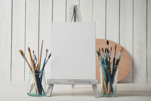 istock Easel with blank canvas and brushes on white wooden table 1322640638