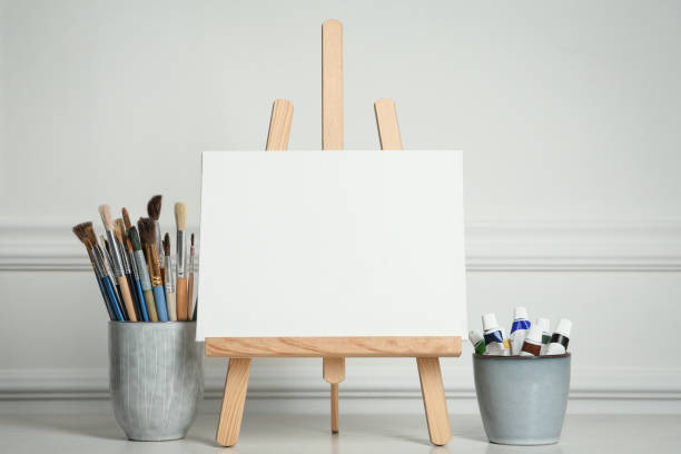 istock Easel with blank canvas, paints and brushes on white wooden table 1322640748
