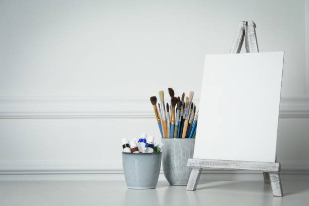 istock Easel with blank canvas, paints and brushes on white wooden table. Space for text 1322640857