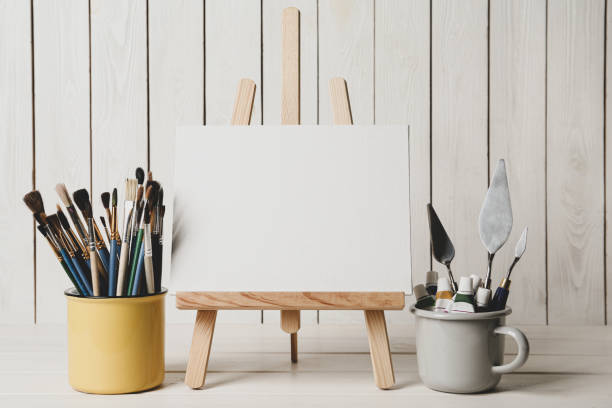 istock Easel with blank canvas, paints and brushes on white wooden table 1322641197