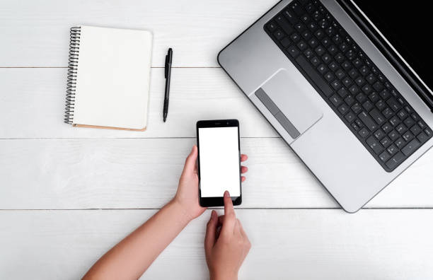 istock Top view on white wooden table with open blank laptop computer, cell phone, empty diary and girl's hands with pen, free space. Girl's hands writing in diary. Mobile phone with white screen copy space 1322643531
