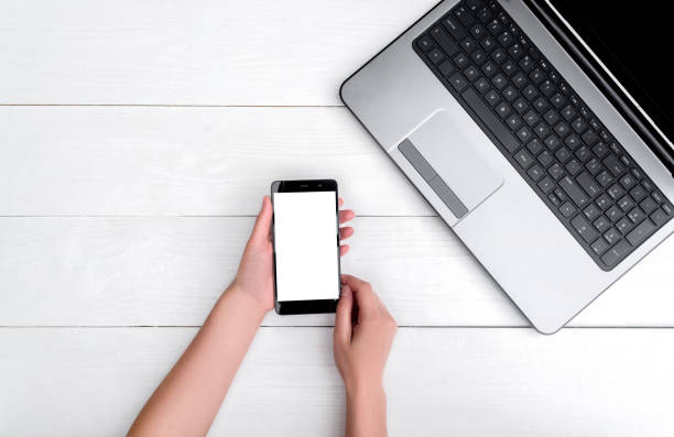istock Top view on white wooden table with open blank laptop computer and girl's hands with cell phone, free space. Mobile phone with white screen, copy space 1322643535