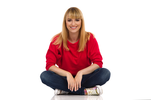 Casual young woman in red sweater sits on a floor and smiles. Front view. Full length studio shot isolated on white.