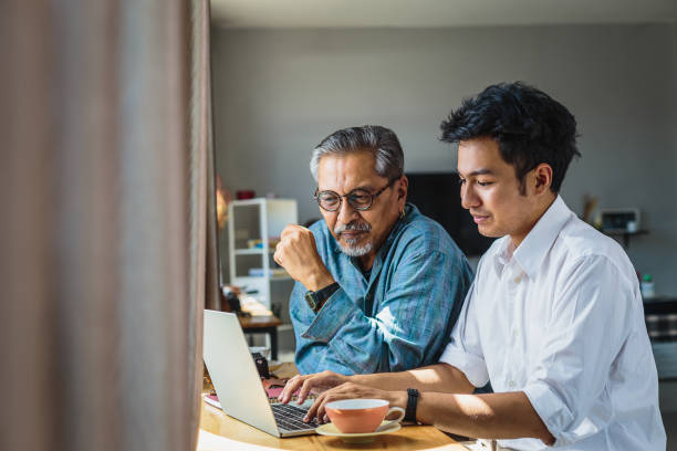 père aîné asiatique et son fils adulte utilisant un ordinateur portable tout en étant assis à la maison - fils de photos et images de collection