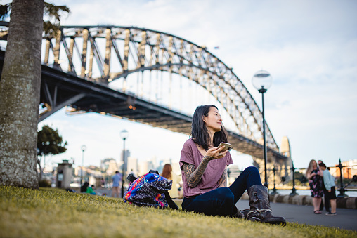 Asian woman in 30's relaxing and using smart phone in Sydney, Australia. Sitting on the grass in the park.
