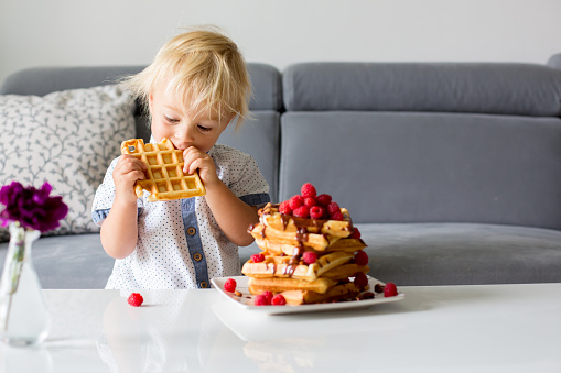 Sweet toddler birthday boy, eating belgian waffle with raspberries and chocolate at home