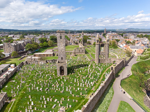 Aerial view of the Royal Burgh of St. Andrews in Fife, located on the east coast of Scotland. The popular university town has a rich cultural past, famous as a place of learning, with a religious history that dates back to the 12th century. The ruin of St. Andrews Cathedral with its ancient cemetery and gravestones can be seen, with its surrounding wall, close to nearby university buildings and other residential homes, seen from a drone view above the town.