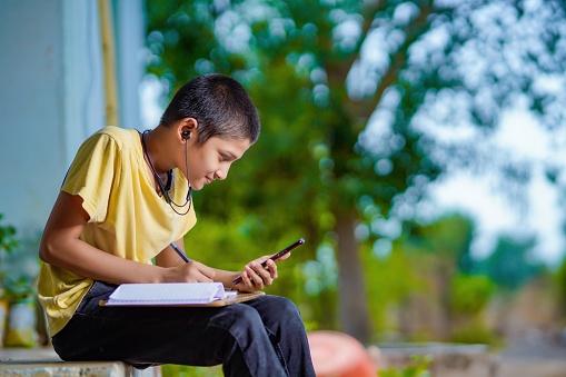 Indian school boy holding phone distance learning class using mobile application, watching online lesson, video calling in app making notes studying at home
