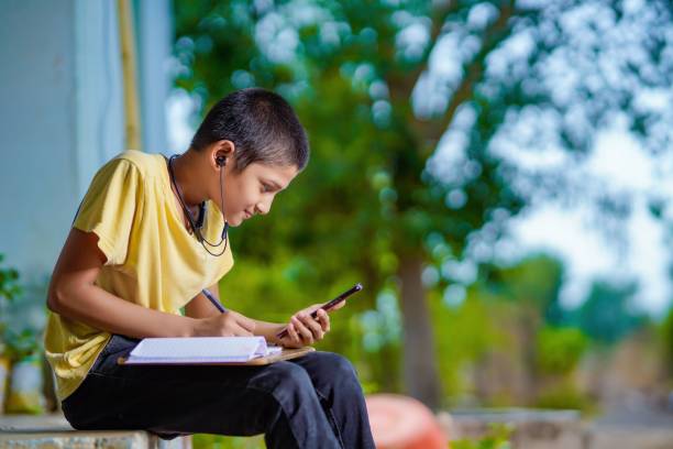 indian school boy holding phone distance learning class using mobile application, watching online lesson, video calling in app making notes studying at home - rural watch imagens e fotografias de stock