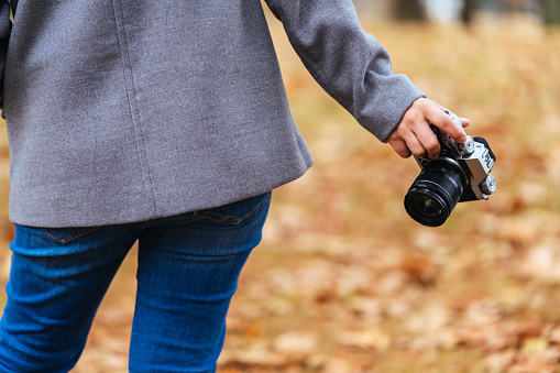 close up female traveller hand hold camera in one hand standing in autumn brown falling tree leaf public nature garden park travel concept