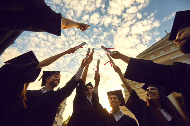 istock Bottom view graduates lift their diplomas and stack them together against a blue sky with clouds. 1322686260