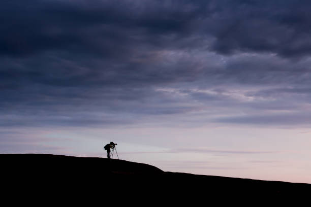 Side view of a young man taking pictures with camera and telephoto zoom lens on tripod on the mountain peak at sunset. Dramatic dark rain clouds in the backgrounds. Travel exploration concepts.