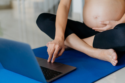 Image of an Asian pregnant woman using laptop for online yoga tutorial. Mother to be using laptop for online exercise class. Home workout, Prenatal care, E-learning.