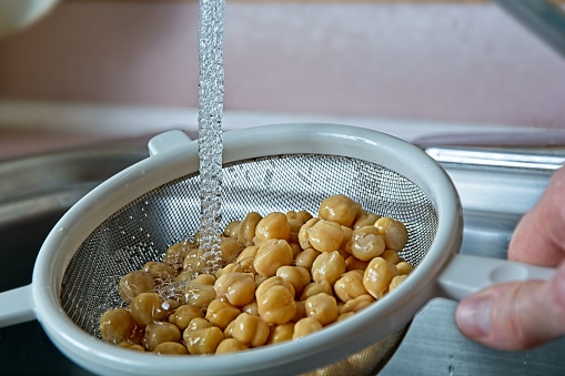 Rinsing chickpeas in a colander under tap water in a stainless steel kitchen sink. The flowing stream of aerated water is frozen to show the glistening bubbles cascading downward  and over the chickpeas below.  The white framed metal strainer is held by hand under the stream of water. Chickpeas are the main ingredient in hummus, a vegetable source of protein and fiber for a healthy diet.
