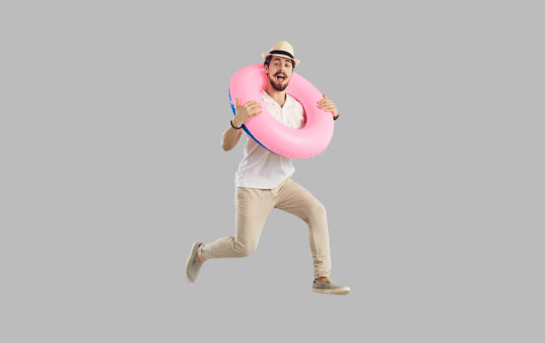 Cheerful traveler tourist man jumping with inflatable ring studio shot portrait Vacation on resort, summer beach rest. Cheerful surprised traveler amazed tourist excited man in casual summer clothes jumping holding inflatable ring. Studio full length portrait over grey copy space people jumping sea beach stock pictures, royalty-free photos & images