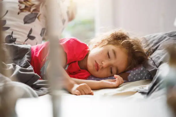 Photo of A small child sleeps sweetly in his bed. Healthy sleep of the girl, ventilation. Morning, wake up, sunlight from the window. Early ascent to kindergarten, school.