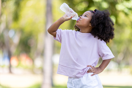 Little African American curly-haired girl concept holding plastic bottles and looking and drinking water on a hot day in the garden.