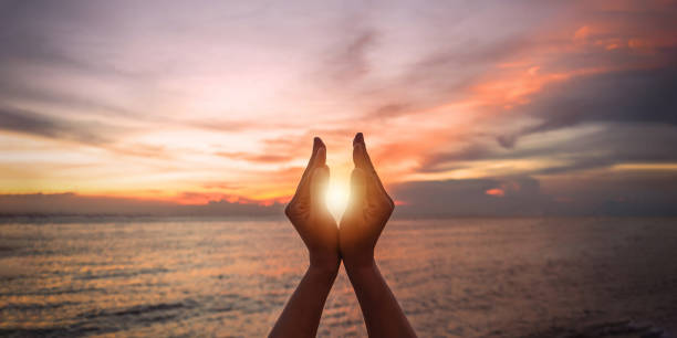 conceito de sol do sol de verão de junho com silhueta das mãos de uma jovem feliz relaxando, meditando e segurando o pôr do sol contra o céu quente da hora dourada na praia com oceano natural ou fundo do mar - heaven light day god - fotografias e filmes do acervo